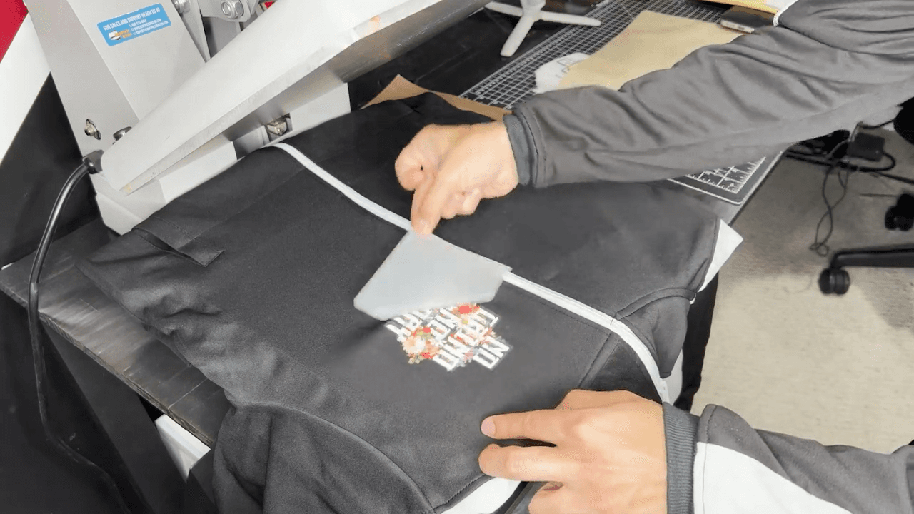Person applying Supacolor transfer design to black track suit using heat press machine